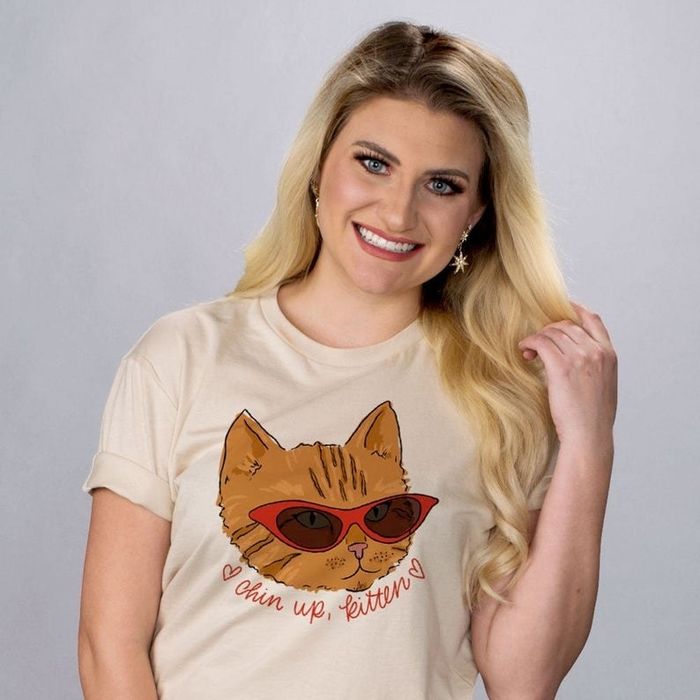 A model wearing a tan tee featuring an orange cat face with red cat-eye sunglasses and the phrase