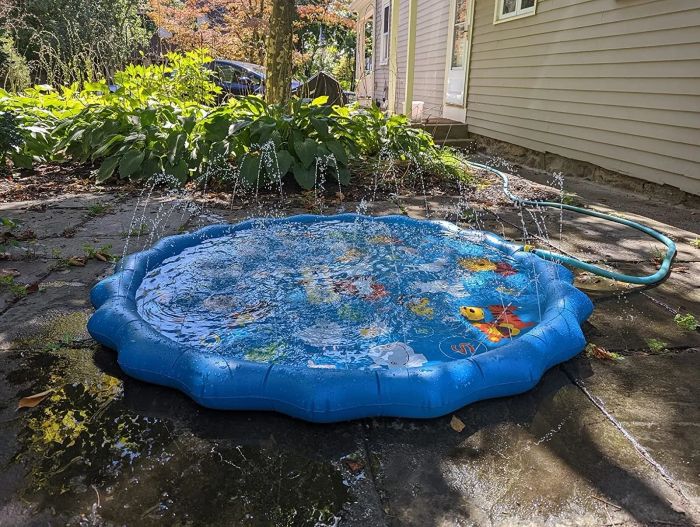 Children's inflatable splash pad featuring a fish design, water sprays along the edges, and a garden backdrop. Perfect for outdoor summer entertainment and playtime