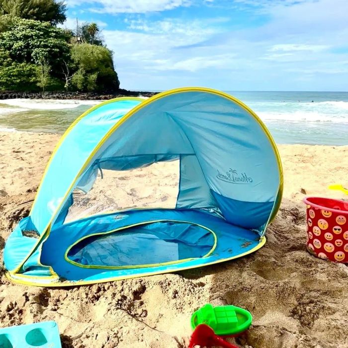 Reviewer's photo of the pop-up beach tent assembled on a sandy beach, with the ocean and trees in the background. A toy bucket, cup, and shovel are visible in the foreground