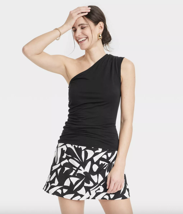 A woman smiling in a black one-shoulder top paired with a patterned skirt, striking a pose with her hand resting on her head