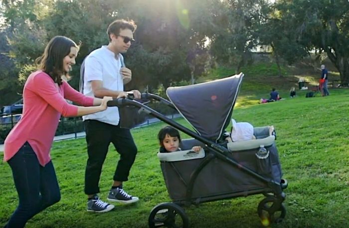 A parent is seen pushing a wagon stroller with a child inside, while another parent walks alongside them in a park setting