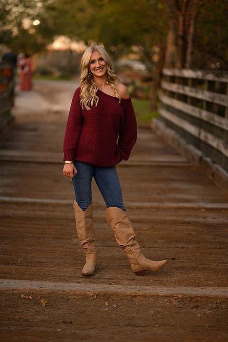 A reviewer wearing a red off-the-shoulder slouchy sweater paired with jeans and knee-high brown boots