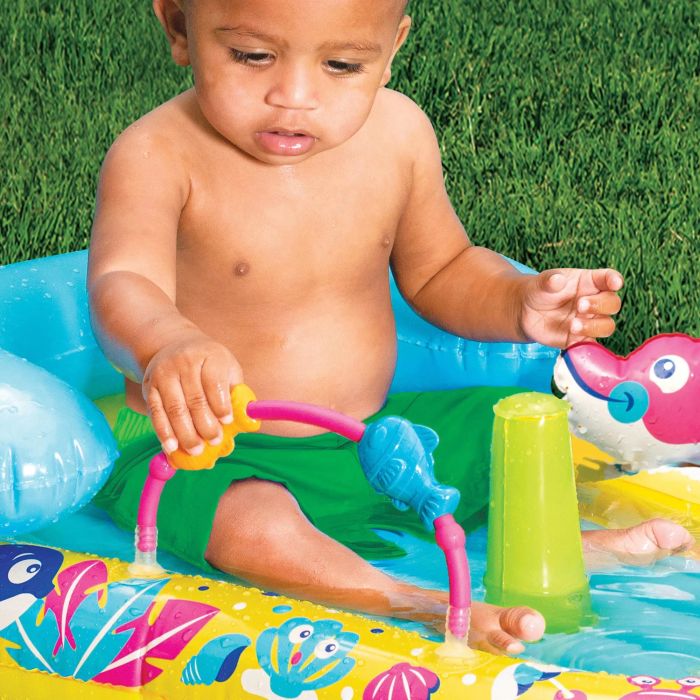A baby in green shorts sits in the splash discovery center, engrossed in playing with a water toy