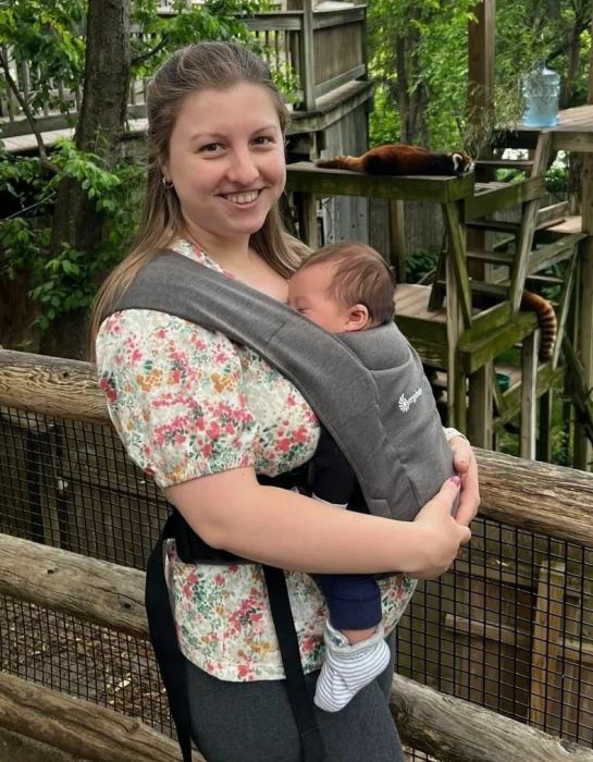 A reviewer in a floral top smiles while holding a baby in a front carrier, with red pandas visible in the background