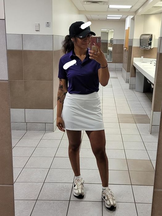 Reviewer taking a restroom mirror selfie, wearing a visor, polo shirt, white skirt, and sneakers
