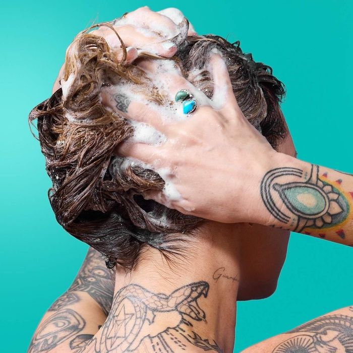 A tattooed individual washing their hair, covered in foamy shampoo, against a solid turquoise backdrop