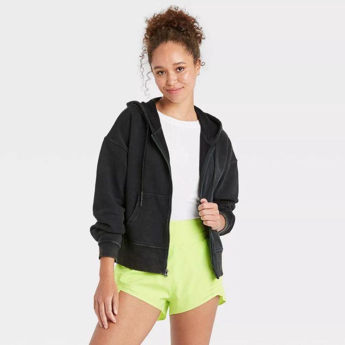 A model wearing the black jacket layered over a white shirt and paired with neon green shorts