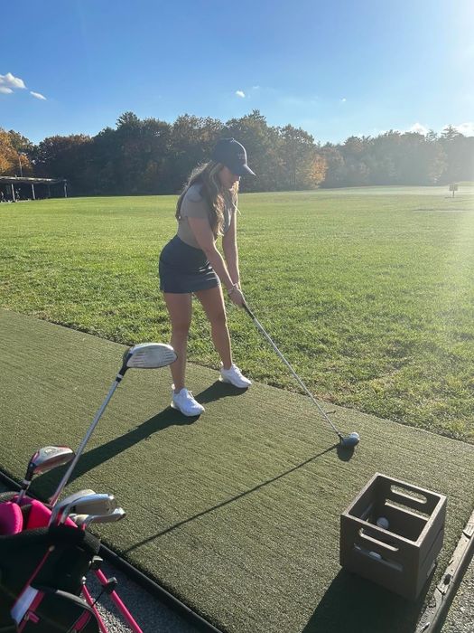 Model preparing to swing at a golf ball on a tee at a driving range, with a golf bag nearby, dressed in appropriate golf attire