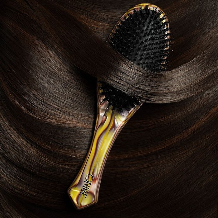 An Oribe hairbrush placed on glossy brown hair, highlighting its sleek design and suitability for premium hair care routines