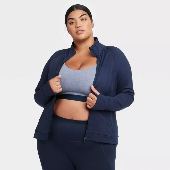 The model is wearing the navy jacket unzipped, paired with a light blue sports bra and navy leggings.
