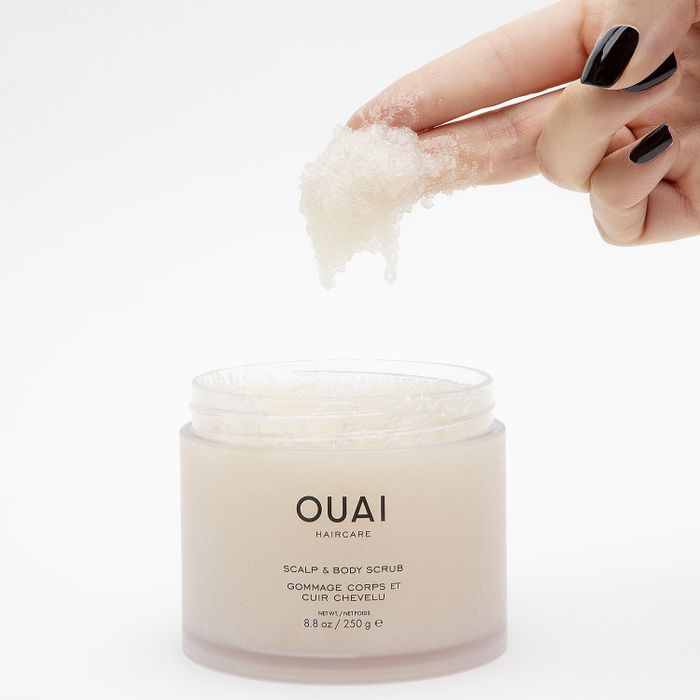 A person’s hand holding a jar of Ouai Scalp & Body Scrub, showcasing the product above its open container.