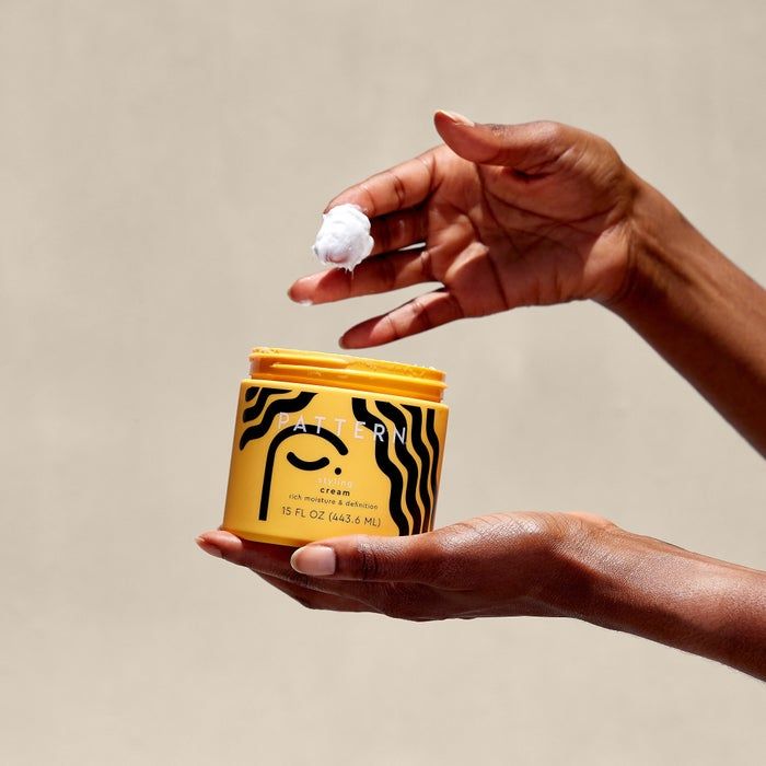 A person holding a jar of Pattern hair cream, demonstrating its smooth texture by scooping a small amount onto their fingers, set against a plain background