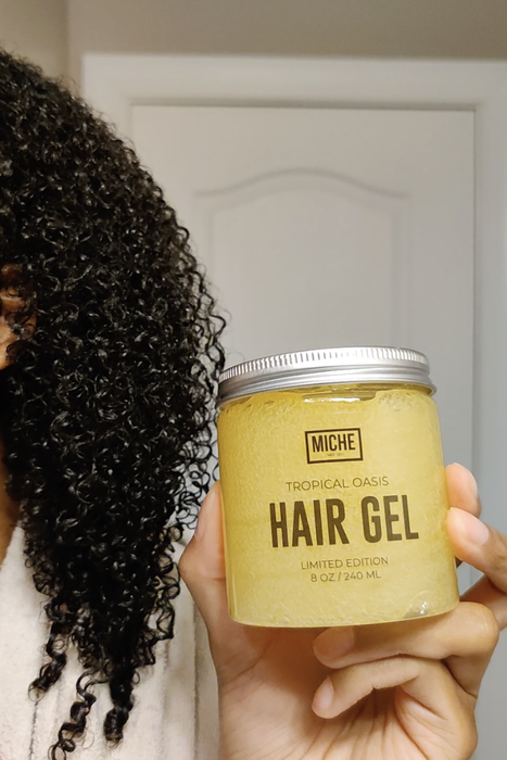 A person holding a jar of Tropical Oasis Hair Gel in front of their curly hair, partially covering their face.