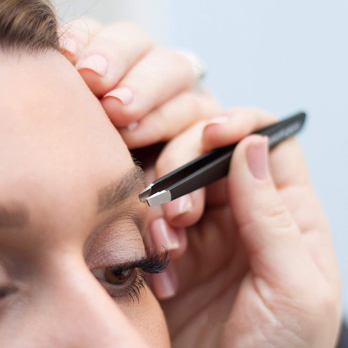A person using slant-tip tweezers to tidy up the hair around their eyebrows.