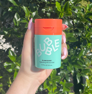 A hand holding the Bubble Slam Dunk Hydrating Moisturizer, 1.7 fl oz, against a backdrop of lush green foliage