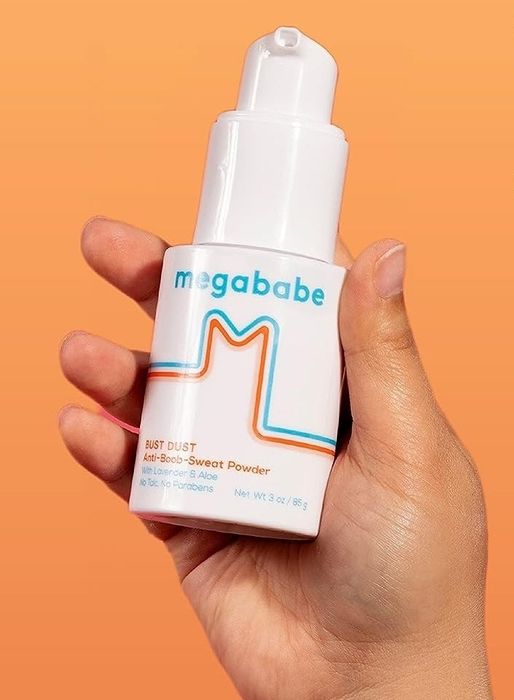 A hand holding a Megababe product container. The label highlights its features: free of talc, synthetic fragrances, aluminum, sulfates, phthalates, and parabens
