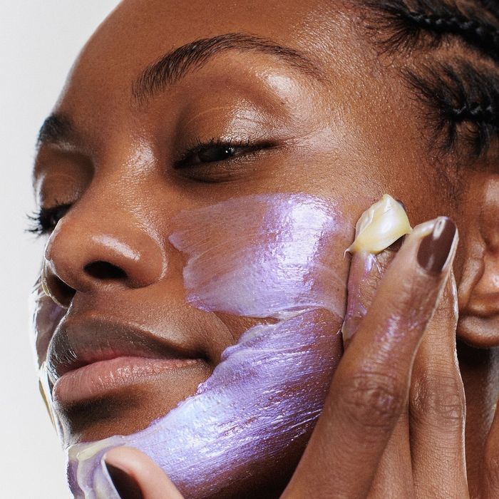 A close-up of someone gently applying a purple cream to their cheek, their expression calm and focused