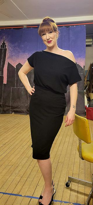 The reviewer is wearing a sleek black off-the-shoulder bodycon dress that ends just at the knee, paired with black heels, standing on a hardwood floor.