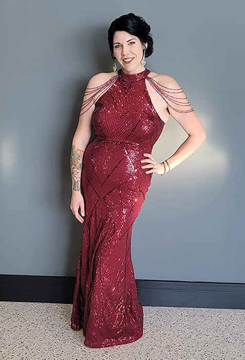 A reviewer in a red sequin maxi halter dress with off-the-shoulder tassels posing against a blue wall