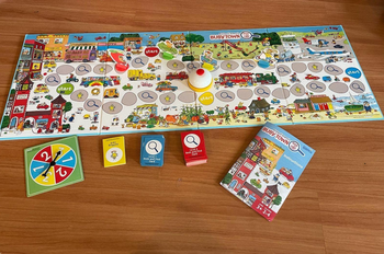 A children's board game set up on a wooden floor, featuring a bright game board, spinner, cards, and an instruction manual, all based on a busy-town theme.