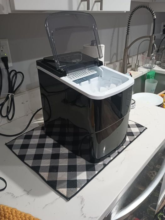 An ice maker with a transparent open lid, placed on a checkered mat on a kitchen counter