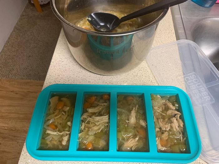 Meal prep featuring chicken soup portioned into a blue tray, placed next to a stainless steel cooking pot