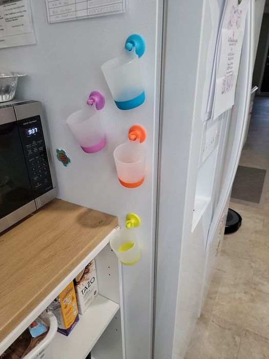 Colorful plastic cups hanging on the side of a refrigerator using hooks, with a microwave and assorted food items visible on the counter