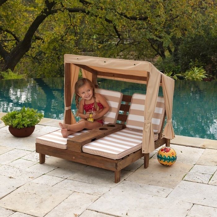 A child relaxes on an outdoor wooden chaise lounge with a canopy by the pool, sipping on a refreshing drink.