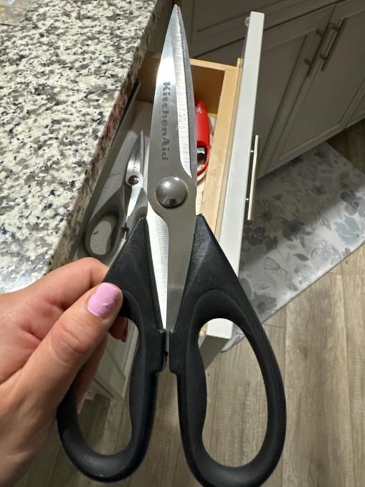 A person holding black-handled KitchenAid kitchen shears above a drawer displaying a red-handled utensil