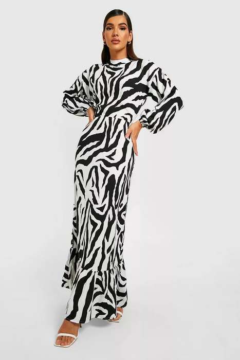 The model is wearing a black and white zebra print maxi dress with balloon sleeves and a high neckline, paired with white open-toe heels against a white backdrop.
