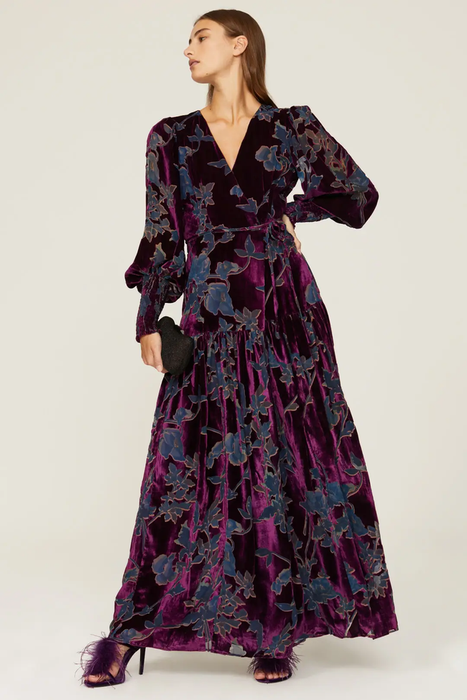 The model is showcasing a purple and navy blue floral velvet maxi dress with a V-neckline and balloon sleeves, styled with feathered purple heels, set against a clean white backdrop.
