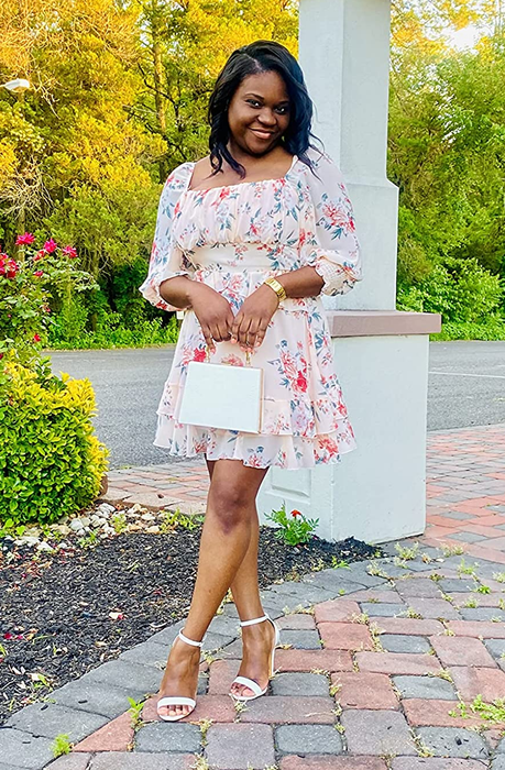 The reviewer is wearing a white floral thigh-length dress with a cinched waist, balloon sleeves, and a ruffled hem, paired with white open-toe heels outdoors.