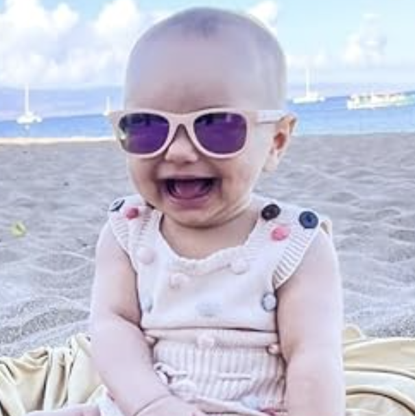 A baby, sporting pink sunglasses and a sleeveless outfit with bright buttons, sits on a beach blanket, smiling at the ocean with sailboats in the distance.