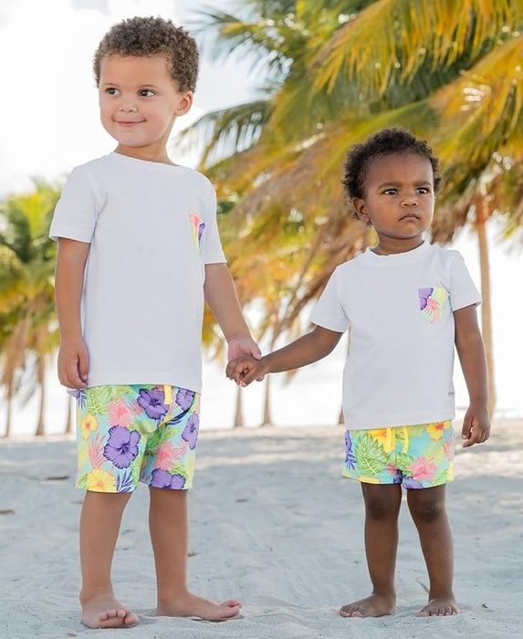 Two young kids holding hands on a beach, wearing white T-shirts and vibrant floral shorts, with palm trees in the background
