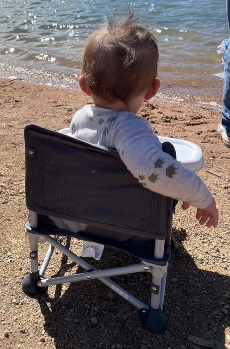 A toddler in a portable chair near a lake, with a dock in the background, and an adult's leg beside them for safety.