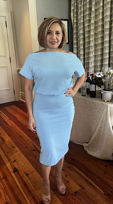 The reviewer is wearing the dress in a soft light blue shade with nude heels, posing on a dark hardwood floor.