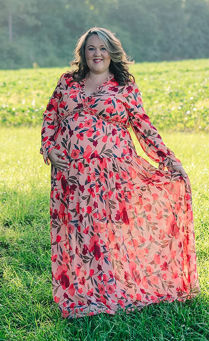 The reviewer is wearing a long-sleeve maxi dress with a pink floral pattern and a ruffled neckline, standing on grass outdoors.