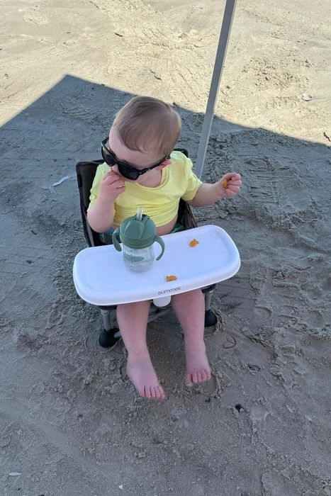 A toddler sits in a high chair on the beach, sporting sunglasses and sipping from a cup.