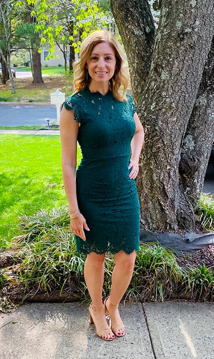 A reviewer looks elegant in a knee-length green lace dress with a crew neckline, paired with nude open-toe heels, standing outdoors in front of a tree.