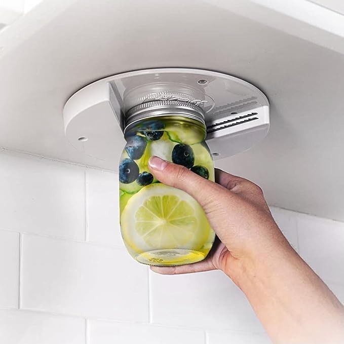 A person attaches a jar filled with water, lime, and blueberries to a white dispenser mounted under a cabinet for easy access