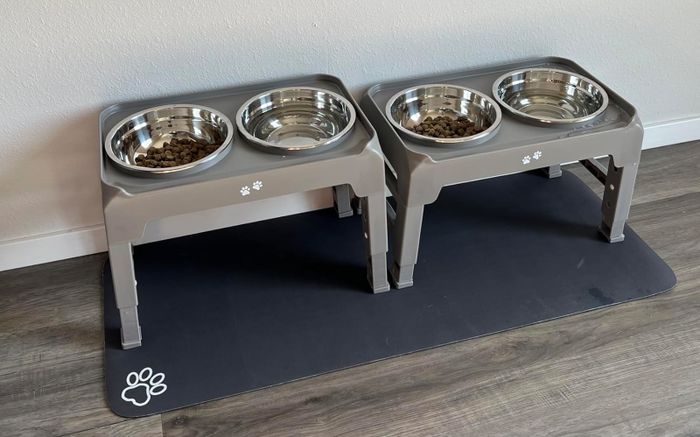 A pair of raised pet food and water bowls positioned on a black mat featuring a paw print design.