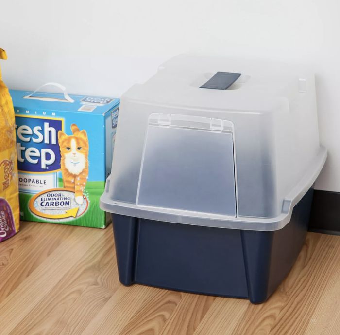 A cat litter box with a cover, placed next to a box of Fresh Step litter and a food bag on a wooden floor.