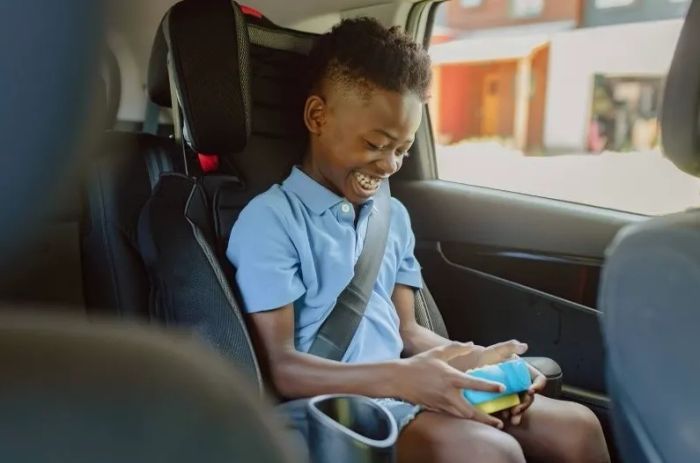 A smiling child seated in a car seat, engrossed in playing with a smartphone