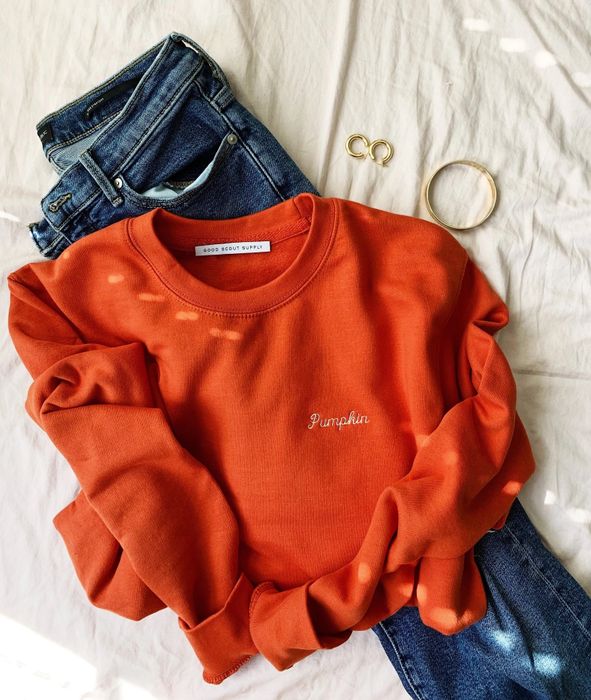 the orange sweatshirt placed on top of a pair of jeans, with some jewelry nearby