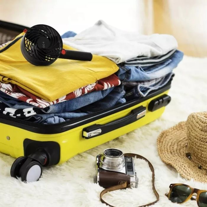 An open suitcase filled with clothes, a hairdryer, a camera, and headphones, alongside a hat and sunglasses, showcasing essential travel items