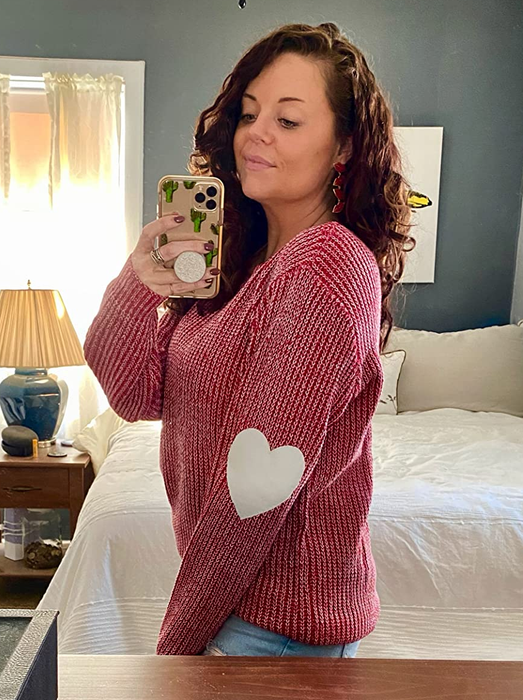 A reviewer capturing a mirror selfie in a cozy bedroom, wearing a red sweater adorned with heart-shaped elbow patches