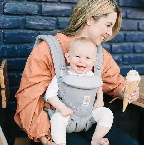 A smiling woman seated with a baby in a front carrier, both facing the camera, as the woman holds an ice cream cone