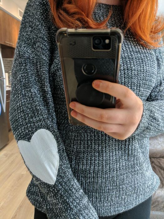 Person snapping a mirror selfie in a gray knitted sweater featuring a heart-shaped elbow patch