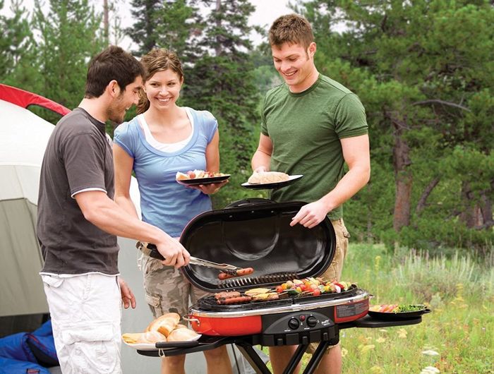three people cooking kabobs and hot dogs on a small portable grill next to a camping tent