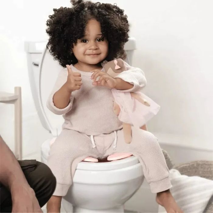 A child sitting on a potty training seat, holding a doll and giving a thumbs up, with an adult’s leg visible nearby